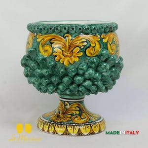 May include: A green and yellow ceramic planter shaped like a pine cone with a decorative base. The planter has a detailed floral design and is made in Italy.