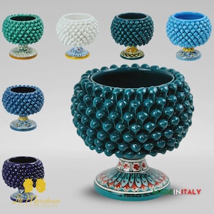 May include: A selection of ceramic vases in shades of teal, white, blue, and purple. Each vase features a textured, pinecone-like pattern and rests on a decorative base with floral designs. The vases are presented against a neutral backdrop.