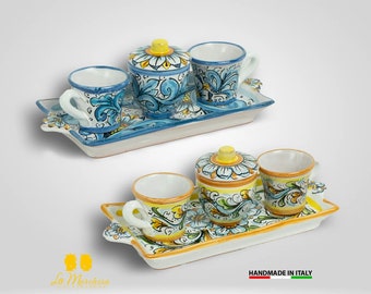 Op de afbeelding: Twee sets Italiaanse keramische espressokopjes en schoteltjes met een bijpassende suikerpot. De kopjes en schoteltjes hebben een blauw en wit bloemenmotief, terwijl de suikerpot een geel en wit bloemenmotief heeft. De sets worden gepresenteerd op bijpassende keramische dienbladen.