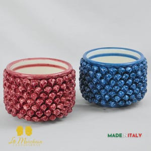Op de afbeelding: Twee keramische plantenbakken, één rode en één blauwe, met een textuur in de vorm van een hart. De plantenbakken hebben een wit interieur en zijn gemarkeerd met "MADE IN ITALY". De plantenbakken zijn waarschijnlijk voor de interieurdecoratie.