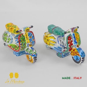 May include: Two ceramic scooter ornaments. The scooters are hand-painted with colourful floral and geometric designs in yellow, blue, green, and red. The scooters have a vintage aesthetic and are set against a white background.