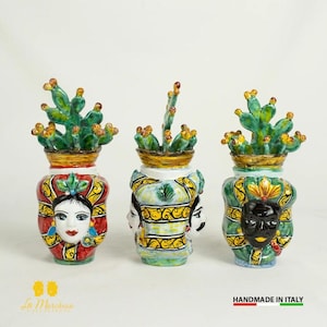 Può includere: Tre vasi in ceramica con volti dipinti e piante di cactus verdi in cima. I vasi sono decorati con motivi e disegni colorati.