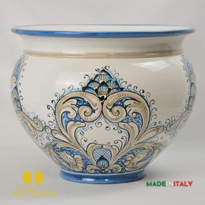 Op de afbeelding: Een grote, ronde keramische plantenbak met een brede opening en een blauwe rand. De gebroken witte body is versierd met een symmetrisch blauw en bruin bloemmotief. De basis is ook blauw en de woorden "MADE IN ITALY" zijn zichtbaar.