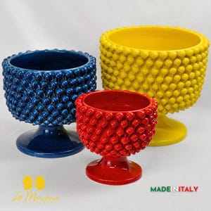 Pode incluir: Três vasos de cerâmica em tamanhos diferentes, azul, vermelho e amarelo, com uma superfície texturizada que se assemelha a uma pinha. Os vasos têm uma base de pedestal.