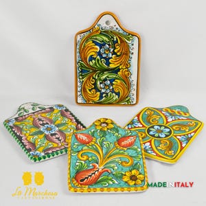 Pode incluir: Quatro bases de cerâmica pintadas à mão com desenhos florais. As bases têm várias formas, incluindo um retângulo e três formas geométricas. A paleta de cores inclui azul, amarelo, verde e laranja. A inscrição "Made in Italy" é visível.