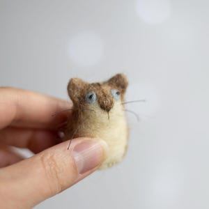 May include: A small needle-felted cat figurine with a brown and cream-coloured coat, light blue eyes, and thin dark whiskers. The text "Felt Art By Mariana" is visible in the upper left corner of the image, featuring a soft, textured wool finish.