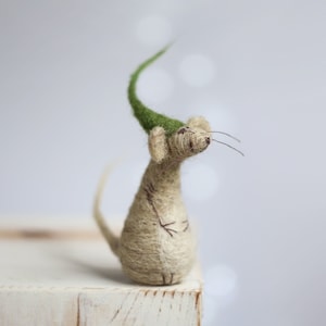 以下が含まれることがあります： 白い木製のブロックの上に、緑色のフェルトの帽子をかぶった、ベージュのフェルトの小さなネズミが座っています。ネズミは上を見上げていて、長い尻尾があります。