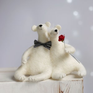 May include: Two white felt polar bears, one wearing a black and white bow tie, are sitting together on a white wooden surface. The bear on the right is holding a red felt rose.