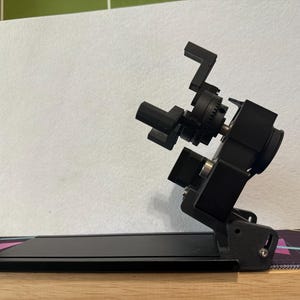 May include: A black, adjustable camera mount with multiple articulating arms and a base. The mount is designed to hold a camera and position it at various angles. The background is a white surface.