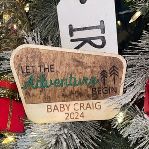 May include: Wooden Christmas ornament with the text "Let the Adventure Begin" and "Baby Craig 2024". The ornament has a wood grain background with green lettering and a white tag. The ornament is hanging on a Christmas tree.