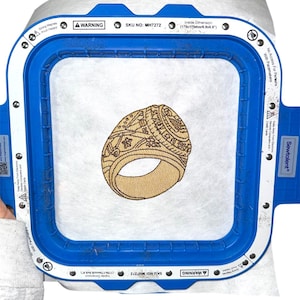 May include: A blue Sewtalent magnetic embroidery hoop with a 6.9 x 6.9 inch inside dimension, holding a white stabilizer sheet featuring a tan-colored embroidered design of a large class ring with detailed star and line patterns.