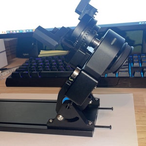 May include: A black camera mount with adjustable arms and a base. The mount is on a wooden surface.