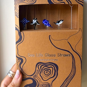 Op de afbeelding: Een bruine doos met een blauw golfpatroon en de tekst "Sea Life Glass Straws". De doos bevat vier glazen rietjes, elk met een zeedierontwerp: een blauwe rog, een zwart-witte rog, een blauwe vis en een zwart-witte orka.