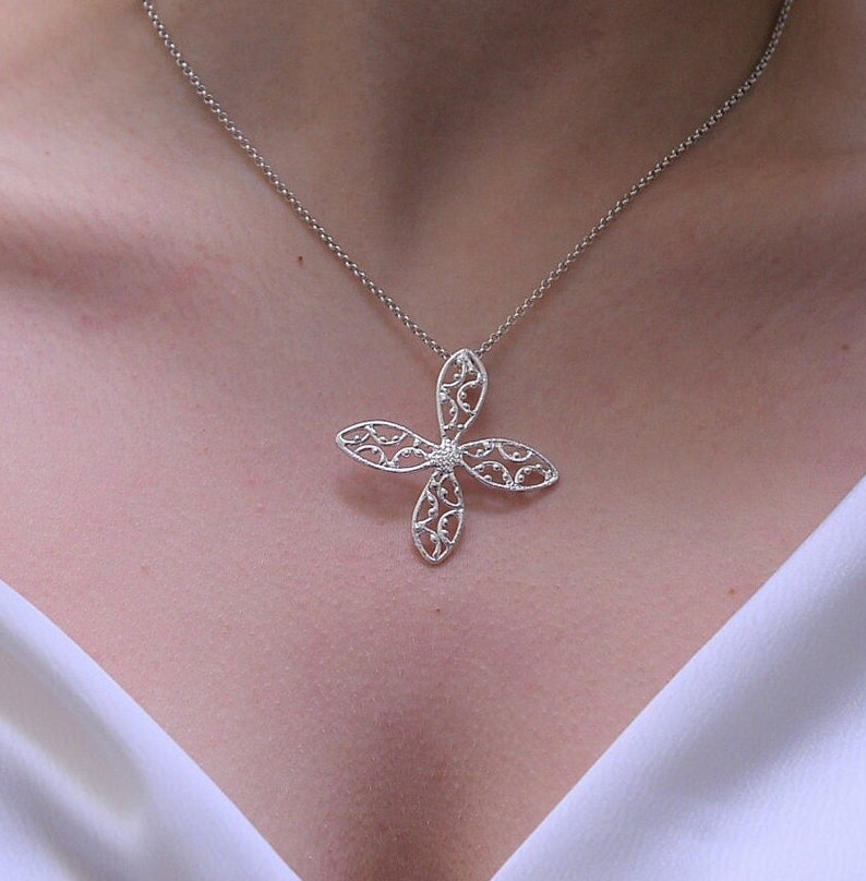 以下が含まれることがあります： 繊細な花の形をしたペンダントが付いたシルバーネックレス。ペンダントは複雑なフィリグリーワークで作られており、中央に小さな透明な石が埋め込まれています。