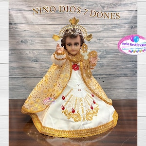 Op de afbeelding: Een gedetailleerd beeldje van Niño Dios, versierd met een gouden kroon en een goudkleurige jurk. De witte en gouden jurk heeft geborduurde details en de tekst "NINO DIOS 7 DONES". Een decoratief religieus item.