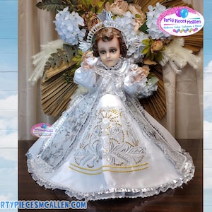 May include: A white and silver dress with gold embroidery and a silver trim. The dress is worn by a doll with a crown and a white dove in its hand. The doll has a white and silver halo. The image includes the text "Party Pieces McAllen".
