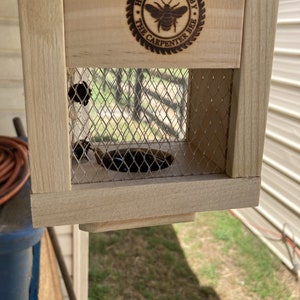 May include: A wooden bee house with a wire mesh opening. The house has a sign that says "Handcrafted by The Carpenter Bee".