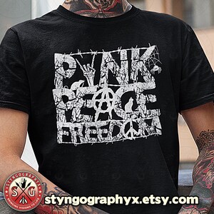 Könnte beinhalten: Schwarzes T-Shirt mit einem weißen Grafikdruck, der "Punk Peace Freedom" mit einem Stacheldraht-Rand sagt. Die Grafik enthält ein Peace-Zeichen, ein Anarchiesymbol und andere Symbole.