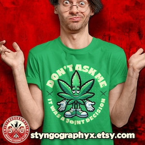Könnte beinhalten: Ein grünes T-Shirt mit einer Cartoon-Marihuana-Blattfigur, die lächelt und die Hände hochhält. Der Text auf dem Shirt lautet "Don't Ask Me It Was A Joint Decision".