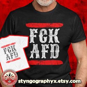 Może przedstawiać: Czarny T-shirt z białym tekstem, który brzmi "FCK AFD" z dwoma czerwonymi liniami nad i pod tekstem. Koszulka jest trzymana przez osobę. Tekst jest w stylu vintage.