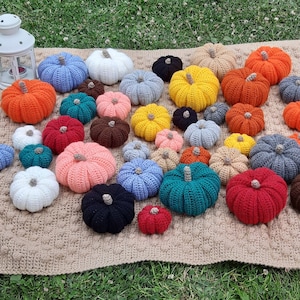 Op de afbeelding: Een verzameling gehaakte pompoenen in verschillende maten en kleuren, waaronder oranje, geel, blauw, wit, roze, rood en bruin. De pompoenen zijn gerangschikt op een getextureerde, beige deken, met een witte lantaarn op de achtergrond. Herfstthema.