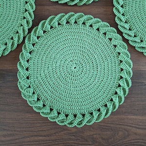 May include: A set of four round, sage green crocheted placemats. Each placemat features a dense, circular center pattern made of tightly worked yarn stitches, surrounded by a decorative, raised, braided-style border along the outer edge.