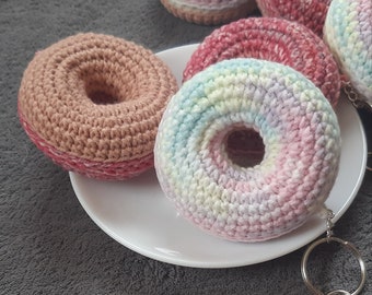 Puede incluir: Cinco llaveros de donut de crochet en varios colores, incluyendo rosa, marrón y arcoíris. Los llaveros están en un plato blanco y una superficie gris.