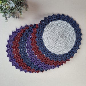 May include: Set of five crocheted placemats in shades of burgundy, purple, and blue. The placemats are round and have a scalloped edge.