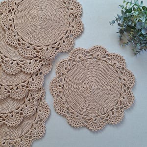 May include: Set of six beige crochet placemats with a delicate lace pattern. The placemats are round and have a scalloped edge.