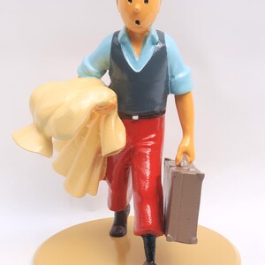 May include: A figurine of a cartoon character with orange hair, wearing a blue shirt, dark vest, and red trousers. The figure is holding a suitcase and a yellow cloth, standing on a tan circular base.