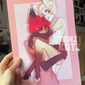 Könnte beinhalten: Ein Druck mit zwei anthropomorphen Figuren in einer Umarmung. Eine Figur hat rotes Haar, die andere hat weißes Fell. Der Druck ist vor einem rosa Hintergrund und in einem passenden rosa Rahmen. Der Text "TANZI KATZ" ist sichtbar.