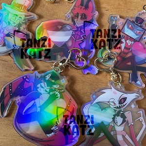 Könnte beinhalten: Eine Sammlung holografischer Acryl-Schlüsselanhänger mit bunten Cartoon-Charakteren. Jeder Schlüsselanhänger hat einen goldfarbenen herzförmigen Verschluss und den Text "TANZI KATZ". Die Charaktere sind in verschiedenen Posen und Designs.