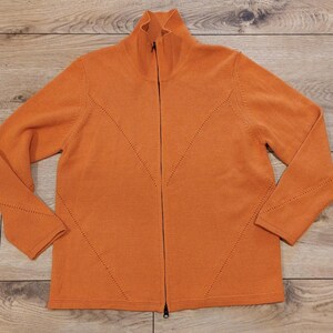 Peut inclure: Une veste zippée en tricot orange uni dotée d'un col montant, de manches longues et d'une fermeture éclair intégrale sur le devant. Le vêtement présente un motif décoratif subtil en forme de V en point ajouré sur la poitrine et les manches.