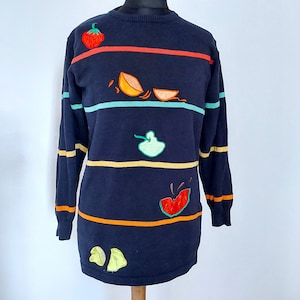 May include: A navy blue striped jumper with colourful embroidered fruit designs, including a strawberry, orange slices, a pear, and a watermelon.