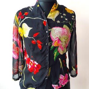 May include: A black sheer blouse with a floral print featuring pink, red, yellow, and green flowers. The blouse has a button-up front and long sleeves.