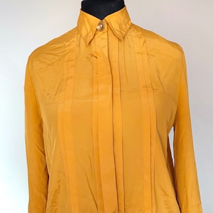 Può includere: Una blusa a maniche lunghe color giallo senape con chiusura a bottoni e design plissettato.