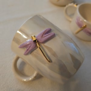 Könnte beinhalten: Weiße Keramiktasse mit einem Libellen-Design in Lila und goldenen Akzenten. Die Libelle hat einen goldenen Körper und lila Flügel.
