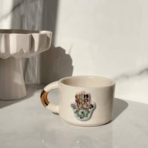 Könnte beinhalten: Eine weiße Keramiktasse mit goldenem Henkel und einem blauen und rosafarbenen Hamsa-Hand-Design. Die Hamsa-Hand ist ein Symbol für Schutz und Glück.