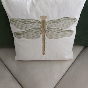 Könnte beinhalten: Ein weißes Zierkissen mit einem gestickten Libellenmuster. Die Libelle ist in Gold- und Olivgrüntönen gehalten. Das Kissen liegt auf einem hellbeigen Sofa, im Hintergrund sind grüne Kissen zu sehen.