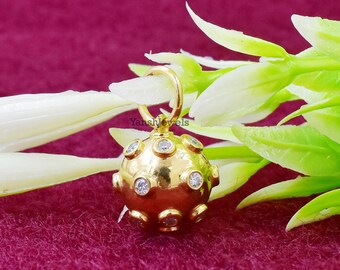 Pode incluir: Um pingente de ouro em forma de esfera com pedras preciosas transparentes engastadas na superfície. O pingente tem um laço na parte superior para prender a uma corrente.