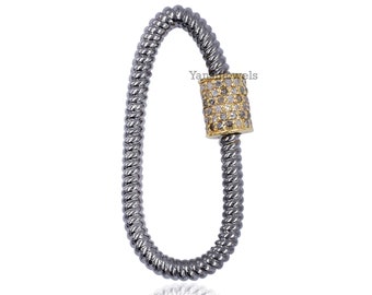 Puede incluir: Una cadena de plata y oro con un dije cilíndrico de diamantes pavé. La cadena está enrollada en forma ovalada.
