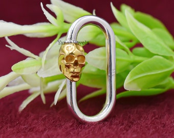 May include: A silver-colored oval-shaped clasp with a gold-colored skull charm. The skull is detailed with eye sockets, a nose cavity, and teeth. The clasp is set against a background of white flowers and green leaves.