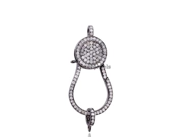 May include: A silver-toned jewelry clasp with a circular top and a teardrop-shaped body. The clasp is encrusted with small, round, clear stones. The clasp is designed to secure jewelry pieces.