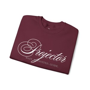 Könnte beinhalten: Burgunderfarbenes T-Shirt mit dem Wort "Projector" in weißer Schrift. Darunter steht der Text "Wait. Be Invited. Guide." Das Shirt ist sauber gefaltet. Der Hintergrund ist weiß.