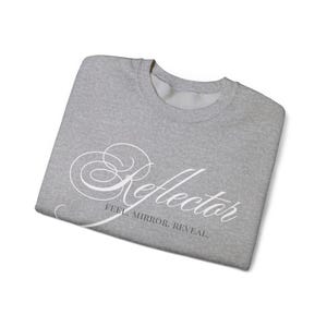 Könnte beinhalten: Ein gefaltetes hellgraues Sweatshirt. Das Wort "Reflector" ist in weißer Schrift aufgedruckt. Darunter steht der Text "FEEL. MIRROR. REVEAL."