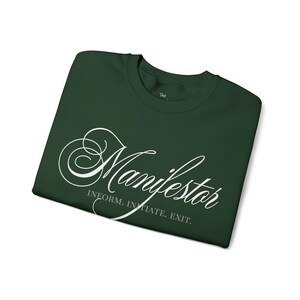 May include: Dark green t-shirt with the word "Manifestor" in elegant white script. Below the word are the words "Inform. Initiate. Exit." The shirt is folded neatly on a white background.