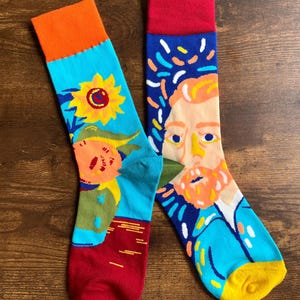 Op de afbeelding: Een paar kleurrijke sokken met artistieke ontwerpen. Eén sok toont een zonnebloem en een figuur, de andere een portret met een blauwe achtergrond en kleurrijke accenten. De sokken hebben oranje, rode, blauwe en gele tinten.