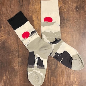 Peut inclure: Une paire de chaussettes de sport avec un motif de paysage. Les chaussettes présentent un soleil rouge, une chaîne de montagnes grise et noire, et un poignet noir. Les chaussettes sont sur une surface en bois.