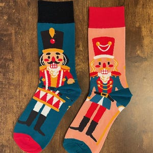 May include: A pair of colourful socks with a nutcracker design. One sock is teal with a red and white drum, the other is pink with a gold and red sceptre. Both socks have a black and white nutcracker illustration.