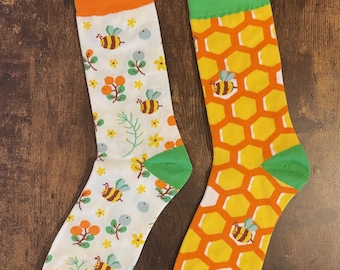 May include: Two pairs of socks with bee-themed designs. One sock features a white background with bees, flowers, and leaves, while the other has a honeycomb pattern. Both socks have orange and green accents.